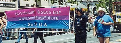 some people marching with a big bi flag stretched on a frame. the flag has "Bi-friendly South Bay - www.southbaybi.org - So B O A" written on it