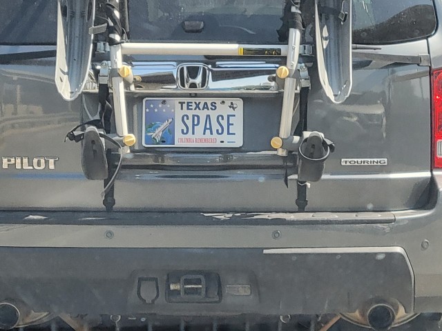 A Texas license plate on a gray Honda Pilot, visible behind a bike rack. The plate's theme is "Columbia remembered." it has a small picture of a rocket with the text to it reading "SPASE" 